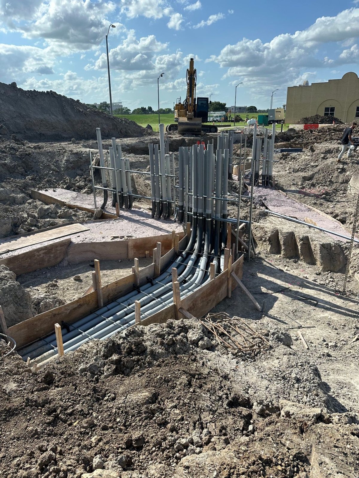PVC conduit riser bundle rising from a concrete-encased duct bank with formwork and excavator on site