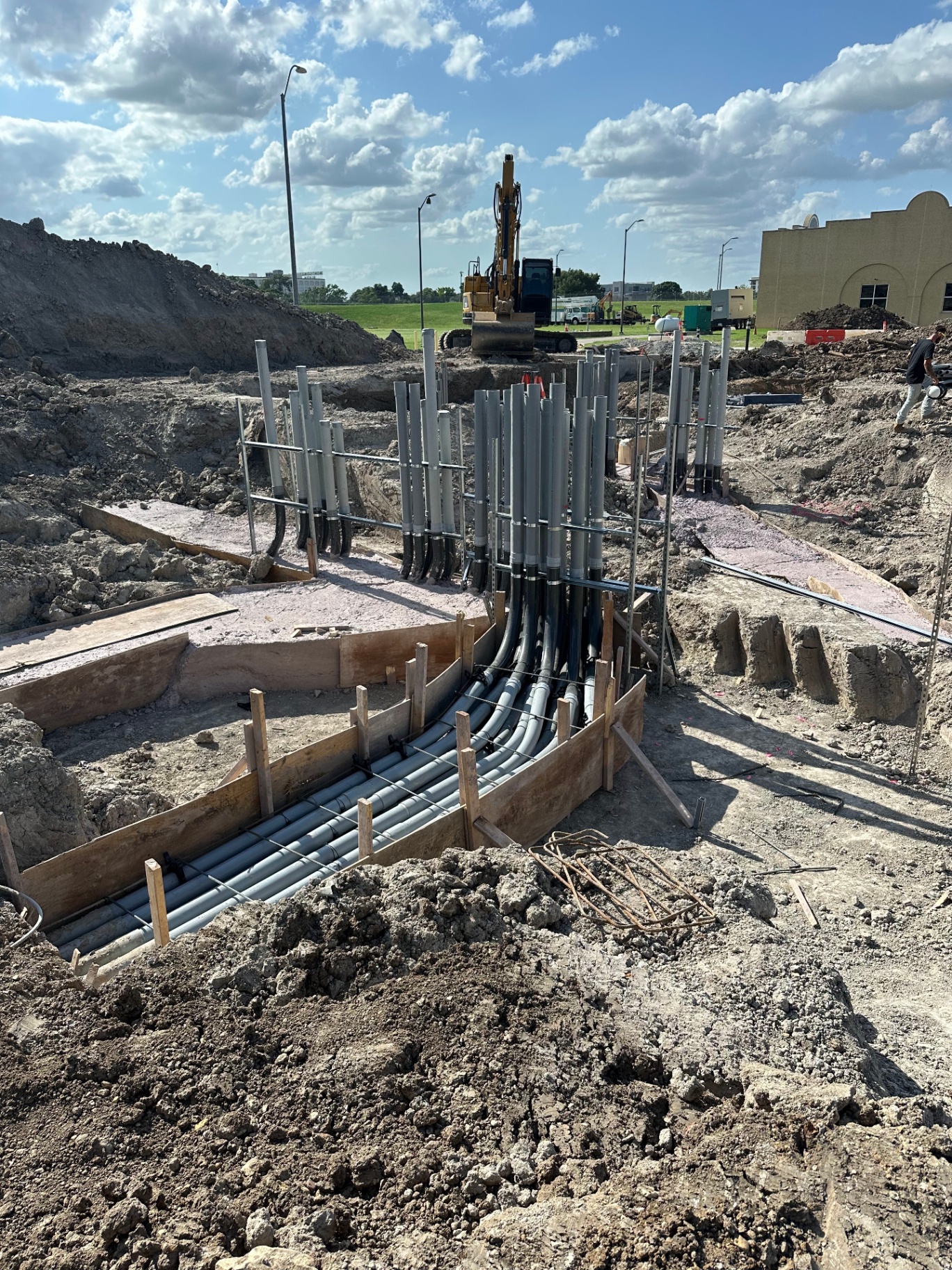 PVC conduit riser bundle rising from a concrete-encased duct bank with formwork and excavator on site