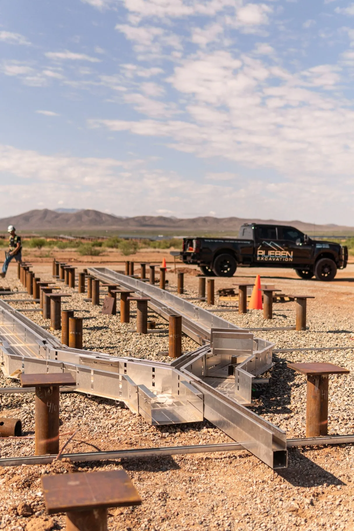 Aluminum cable tray and steel pile caps staged on a gravel jobsite with a Rueben Excavation truck and desert mountains in the background